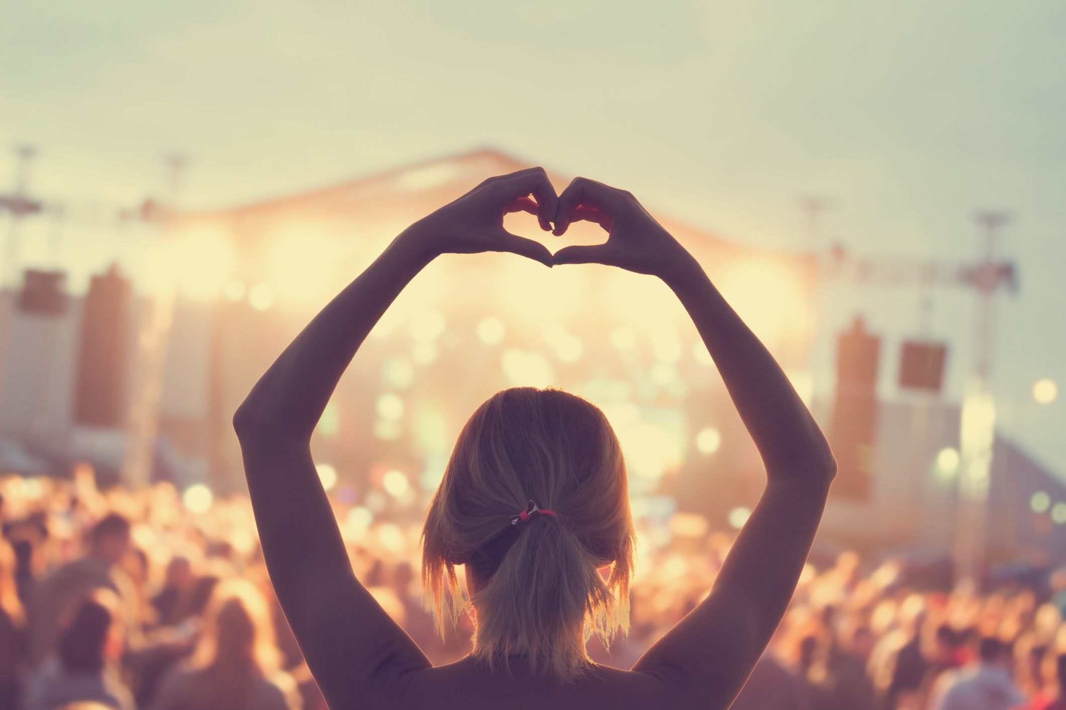A female is listening to music live at a festival in a crown and she is creating a heart shape with her hands